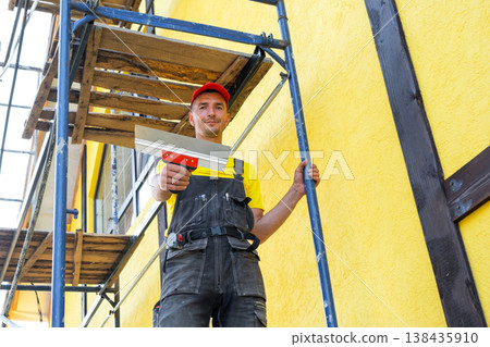 Plasterer with trowel on facade, plastering and smoothing exterior walls, surface leveling, decorative finish, private business, professional wall preparation. 138435910