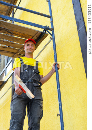 Master plasterer with trowel on scaffolding, facade works, applying and leveling compound, exterior finishing, private renovation business, house improvement. 138435911