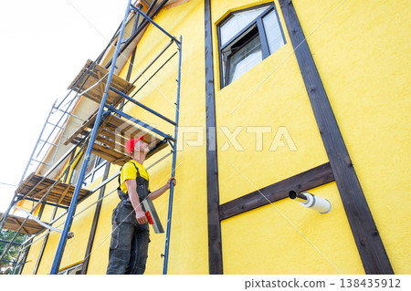 Plasterer on scaffolding working on facade, applying plaster, leveling walls, exterior renovation, decorative finishing, private business, building services. 138435912