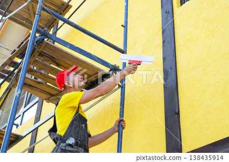 Decorator with trowel on scaffolding, applying decorative plaster, facade smoothing, wall renovation, private business, building exterior preparation service. 138435914
