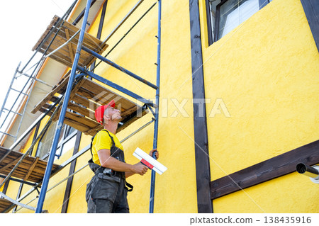 Plasterer craftsman levels facade wall with trowel, applying compound, smoothing, decorative finish, private renovation business, building exterior maintenance. 138435916