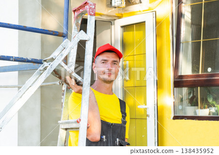 Builder in yellow uniform and red cap with ladder, smiling at camera near home, construction field service business visit, outdoor renovation works. Builder in yellow uniform and red cap with ladder, smiling at camera near home, construction field service business visit, outdoor renovation works. 138435952