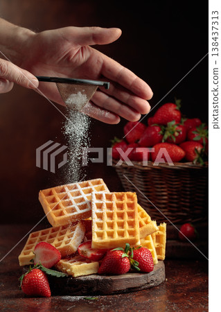 The chef sprinkles powdered sugar on the waffles topped with fresh strawberries. The chef sprinkles powdered sugar on the waffles topped with fresh strawberries. 138437313