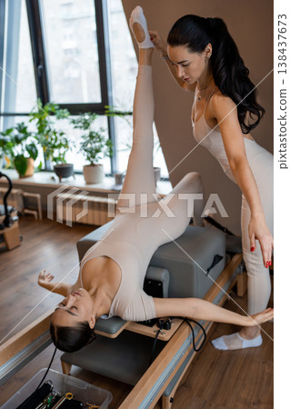 Pilates instructor adjusting foot position while woman holds vertical leg pose on reformer in airy studio. Vertical leg pose, reformer, foot position, studio, instructor. Pilates instructor adjusting foot position while woman holds vertical leg pose on reformer in airy studio. Vertical leg pose, reformer, foot position, studio, instructor. 138437673