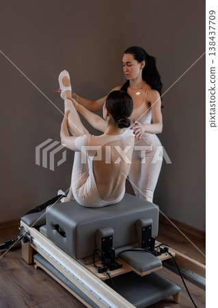 Trainer giving assisted stretch to client's legs on pilates reformer during advanced pilates session. Assisted stretch, pilates reformer, trainer, client, advanced. Trainer giving assisted stretch to client's legs on pilates reformer during advanced pilates session. Assisted stretch, pilates reformer, trainer, client, advanced. 138437709