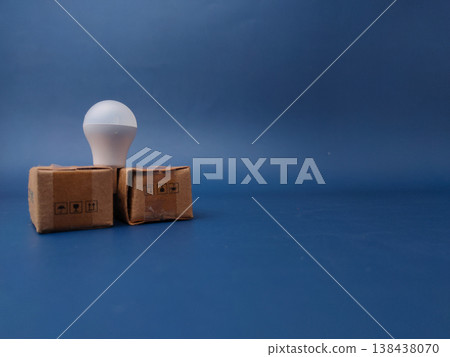 A minimalist studio shot featuring a small white USB LED light bulb balanced between two brown cardboard shipping boxes against a solid blue background. A minimalist studio shot featuring a small white USB LED light bulb balanced between two brown cardboard shipping boxes against a solid blue background. 138438070