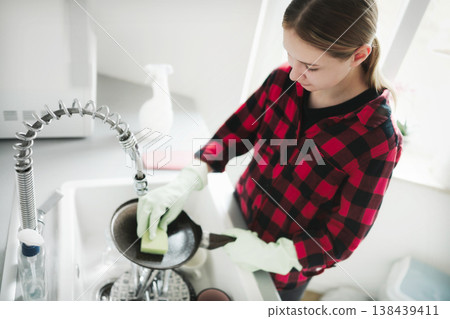 Teenage girl washes dishes in a modern kitchen while wearing gloves. Washing a frying pan with a sponge on . Teenage girl washes dishes in a modern kitchen while wearing gloves. Washing a frying pan with a sponge on . 138439411