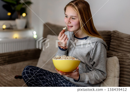 Girl on couch with brown blanket, popcorn in hand, enjoying a movie in a cozy room 138439412