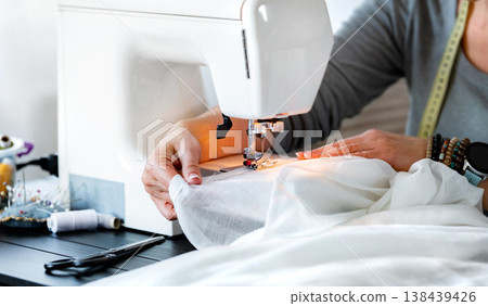 Close up of Female tailor hands sewing fabric in sewing machine. Female worker sewing with machine to hem curtains. DIY process of sewing in home workshop. Close up of Female tailor hands sewing fabric in sewing machine. Female worker sewing with machine to hem curtains. DIY process of sewing in home workshop. 138439426