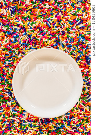 Top-down view of an empty white plate centered on a vibrant background of rainbow-colored sprinkles. 138439602