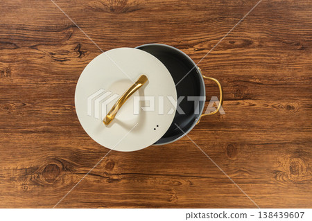 Minimal flat lay of a lidded gray pot with a golden handle centered on a wood table. The lid rests slightly tilted revealing the pot rim. Minimal flat lay of a lidded gray pot with a golden handle centered on a wood table. The lid rests slightly tilted revealing the pot rim. 138439607