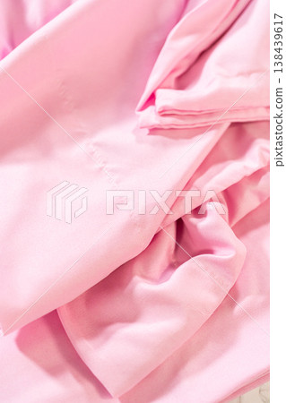 Light pink bed sheet appears partially folded and wrinkled on a light surface. Fabric texture and loose folds are clearly visible. Light pink bed sheet appears partially folded and wrinkled on a light surface. Fabric texture and loose folds are clearly visible. 138439617