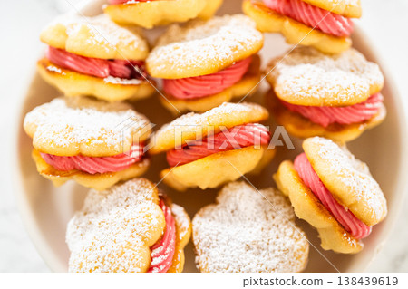 A bowl filled with lemon spritz cookie sandwiches, each piped with vibrant cranberry buttercream and dusted with powdered sugar. The cookies have a delicate, buttery texture with a festive touch A bowl filled with lemon spritz cookie sandwiches, each piped with vibrant cranberry buttercream and dusted with powdered sugar. The cookies have a delicate, buttery texture with a festive touch 138439619