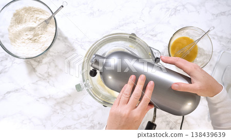 Hands operate a stand mixer blending cookie dough mixture inside a glass bowl on a marble surface. Flour and beaten eggs are arranged nearby, emphasizing organized baking preparation. Bright lighting Hands operate a stand mixer blending cookie dough mixture inside a glass bowl on a marble surface. Flour and beaten eggs are arranged nearby, emphasizing organized baking preparation. Bright lighting 138439639