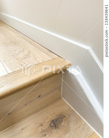 A close up shows a wooden stair step beside a white wall with trim. The detail is clean, minimal, and modern in design. A close up shows a wooden stair step beside a white wall with trim. The detail is clean, minimal, and modern in design. 138439641