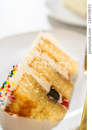 A diagonally angled slice of vanilla sponge cake with blueberry and cream sits on a white plate. The frosting edge is decorated with bright sprinkles. The background is softly blurred to focus on the 138439653