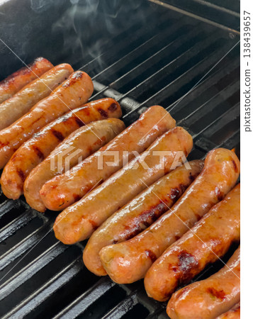 Multiple sausages cook on a grill with visible char and rising smoke. The grill grates are hot and blackened from use. Multiple sausages cook on a grill with visible char and rising smoke. The grill grates are hot and blackened from use. 138439657