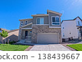 A two-story modern suburban home with a stone accent facade and large garage under clear blue sky. The house is surrounded by neighboring homes and landscaped with grass and a small tree. Captured 138439667