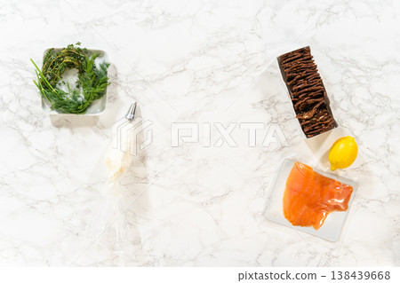 A top view of fresh herbs a piping bag with a metal tip smoked salmon crispbread crackers and a lemon arranged on a marble surface before assembling canapes 138439668