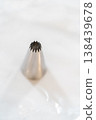 A single metal piping tip is isolated on a white marble surface. Bright lighting highlights clean shape and baking tool detail. 138439678