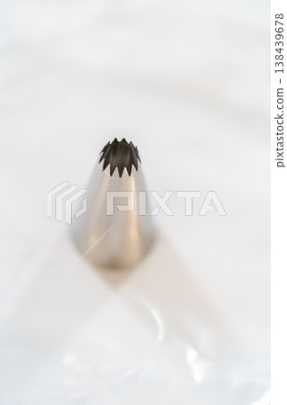 A single metal piping tip is isolated on a white marble surface. Bright lighting highlights clean shape and baking tool detail. A single metal piping tip is isolated on a white marble surface. Bright lighting highlights clean shape and baking tool detail. 138439678