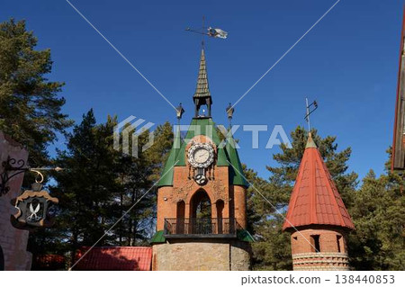 Historic Clock Tower Amid Nature Scenery. Decorative Tower With Spires Under Bright Sky And Pine Trees Historic Clock Tower Amid Nature Scenery. Decorative Tower With Spires Under Bright Sky And Pine Trees 138440853