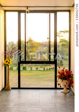 Interior view through large open window looking out to green landscape at sunset. Flower vases with sunflowers and tulips on the floor. Warm golden hour light illuminating the room and garden. Interior view through large open window looking out to green landscape at sunset. Flower vases with sunflowers and tulips on the floor. Warm golden hour light illuminating the room and garden. 138442746