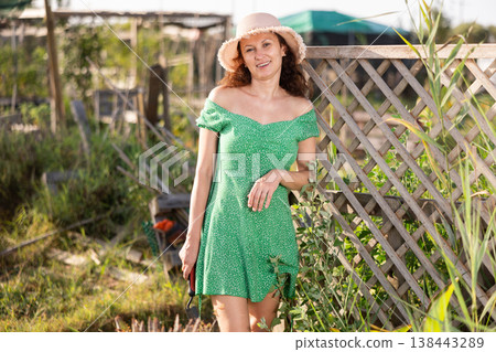 Slender woman in green dress and straw hat poses in her garden, in the backyard of country house Slender woman in green dress and straw hat poses in her garden, in the backyard of country house 138443289