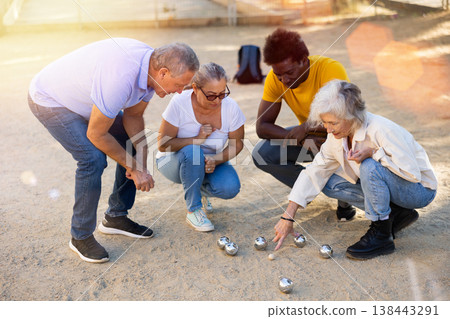 Focused elderly multinational people squatting in a circle and looking at the metal petanque balls on the sandy area at the park Focused elderly multinational people squatting in a circle and looking at the metal petanque balls on the sandy area at the park 138443291