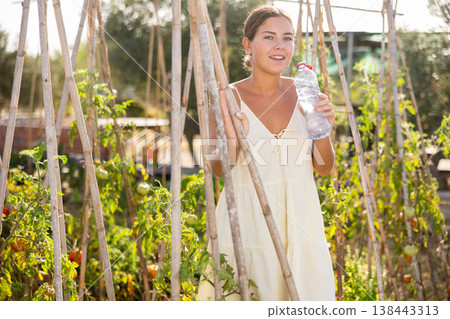 Hot summer weather in the garden beds of a country house - young woman drinks water from a bottle Hot summer weather in the garden beds of a country house - young woman drinks water from a bottle 138443313