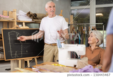 Sewing school senior man teacher conducts lesson for elderly students, draws pattern on black-board Sewing school senior man teacher conducts lesson for elderly students, draws pattern on black-board 138443467