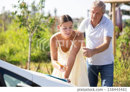 Elderly man shows a young woman where to sign a document. A man and a woman sign documents on the hood of car Elderly man shows a young woman where to sign a document. A man and a woman sign documents on the hood of car 138443481