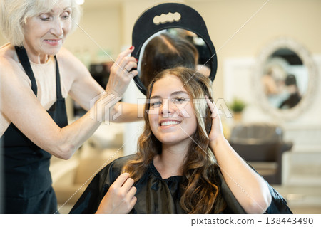 Elderly female hairdresser showing result of hairstyling with mirror to young girl Elderly female hairdresser showing result of hairstyling with mirror to young girl 138443490