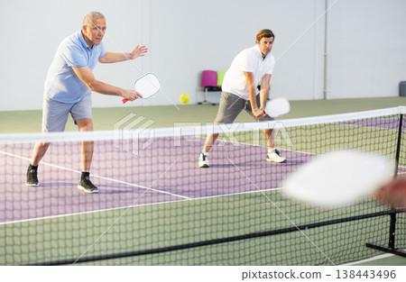 Senior man playing doubles pickleball with male partner indoors 138443496