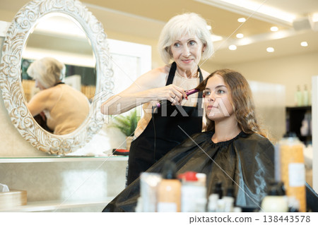 Elderly female hairstylist using curling wands to add volume for hair of young girl 138443578