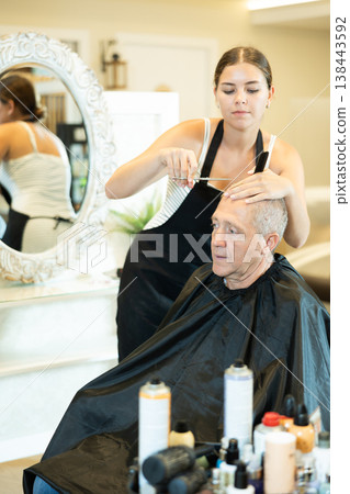 Young girl hairstylist making haircut to elderly man in hairdressing salon Young girl hairstylist making haircut to elderly man in hairdressing salon 138443592