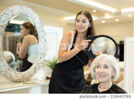 Young hairstylist holding mirror while presenting result of styling to elderly woman Young hairstylist holding mirror while presenting result of styling to elderly woman 138443769