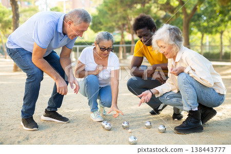 Focused elderly multinational people squatting in a circle and looking at the metal petanque balls on the sandy area at the park 138443777