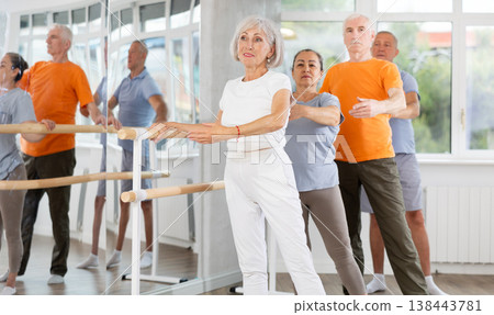 Active mature woman practicing battement tendu holding by ballet barre together with others Active mature woman practicing battement tendu holding by ballet barre together with others 138443781