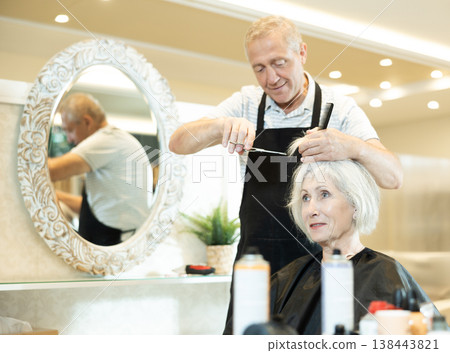 Elderly hairdresser using scissors while providing haircut to woman in salon Elderly hairdresser using scissors while providing haircut to woman in salon 138443821