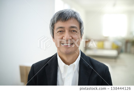 A man in his 60s wearing a jacket and smiling in his room. 138444126