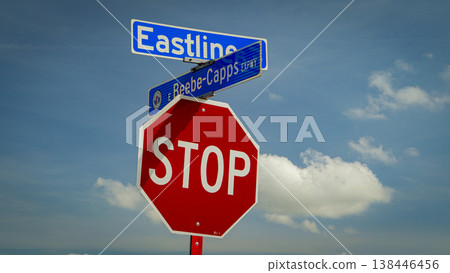 Red octagonal stop sign mounted beneath blue street signs at an intersection against a bright blue sky with fluffy clouds, conveying traffic control, safety, direction and urban signage 138446456