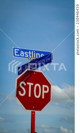Red octagonal stop sign mounted beneath blue street signs at an intersection against a bright blue sky with fluffy clouds, conveying traffic control, safety, direction and urban signage 138446459