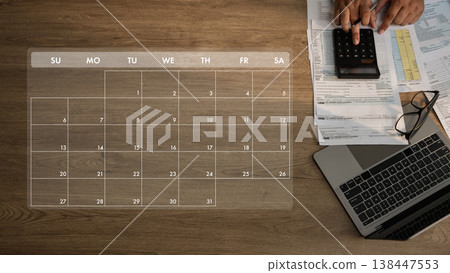 Top view of a person calculating budget planning documents with a calculator on a desk with a laptop and eyeglasses, with a calendar overlay indicating payment schedule and due date planning. Top view of a person calculating budget planning documents with a calculator on a desk with a laptop and eyeglasses, with a calendar overlay indicating payment schedule and due date planning. 138447553