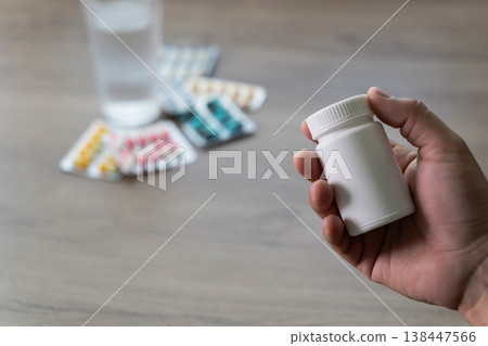 Close up of hand holding a white medicine pill bottle with blister packs and glass of water for healthcare treatment, showing professional pharmacy medication and a beautiful wellness concept Close up of hand holding a white medicine pill bottle with blister packs and glass of water for healthcare treatment, showing professional pharmacy medication and a beautiful wellness concept 138447566