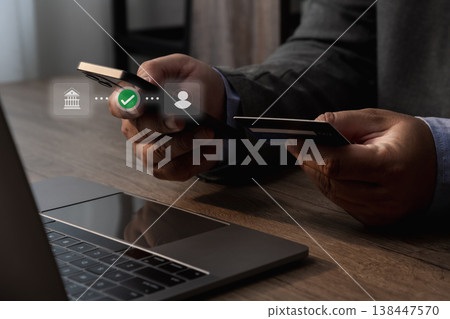 Close-up of hands holding a smartphone and credit card over a laptop, showing mobile payment verification with check icon, representing secure online transaction and digital banking. 138447570