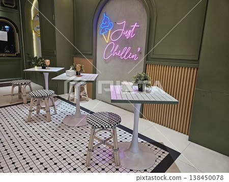 A stylish modern cafe interior featuring olive green walls, elegant checkered flooring, and marble tables illuminated by a vibrant "Just Chillin" pink neon sign with a blue ice cream cone icon. A stylish modern cafe interior featuring olive green walls, elegant checkered flooring, and marble tables illuminated by a vibrant "Just Chillin" pink neon sign with a blue ice cream cone icon. 138450078