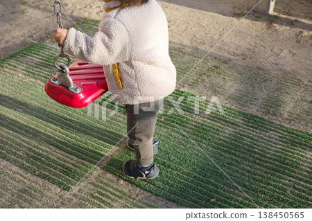 一個穿著冬裝的孩子在公園玩耍，抓著鞦韆的鏈條。 138450565