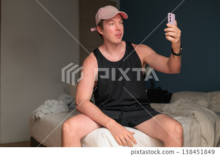 Caucasian Man in Pink Cap Taking Selfie with Smartphone on Bed in Bedroom 138451849