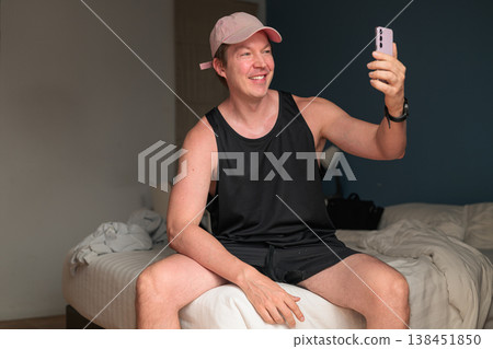Smiling Caucasian Man in Pink Cap Taking Selfie with Smartphone on Bed in Bedroom 138451850